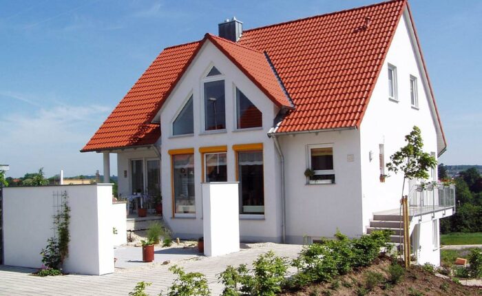 Weißes Einfamilienhaus mit rotem Satteldach, Garten und Terrasse bei sonnigem Wetter.