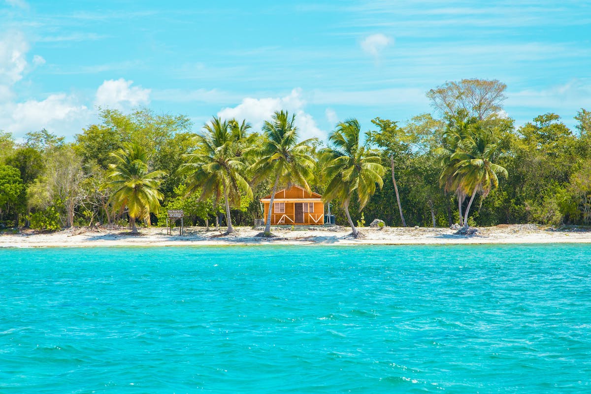 Kleines Holzhaus an einem tropischen Strand, umgeben von Palmen und grünem Dschungel, mit türkisblauem Meer im Vordergrund.