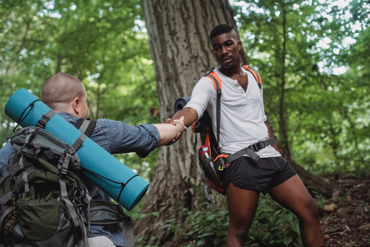 Zwei Wanderer im Wald, einer hilft dem anderen mit ausgestreckter Hand beim Aufstieg, beide mit Rucksäcken und Ausrüstung.